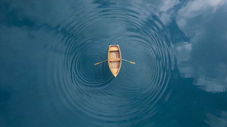 Vertical video: Starting ripples moving outward beneath wooden rowboat at calm blue lake, with oars