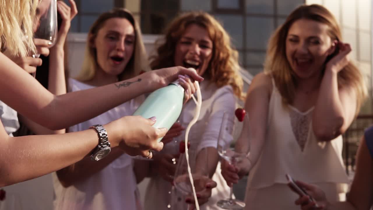 hermosas damas jóvenes celebran. despedida de soltera. sacudiendo una extraña botella de champán y dejándola rociar. la chica vierte una bebida en vasos. afuera. fiesta glamorosa. cámara lenta