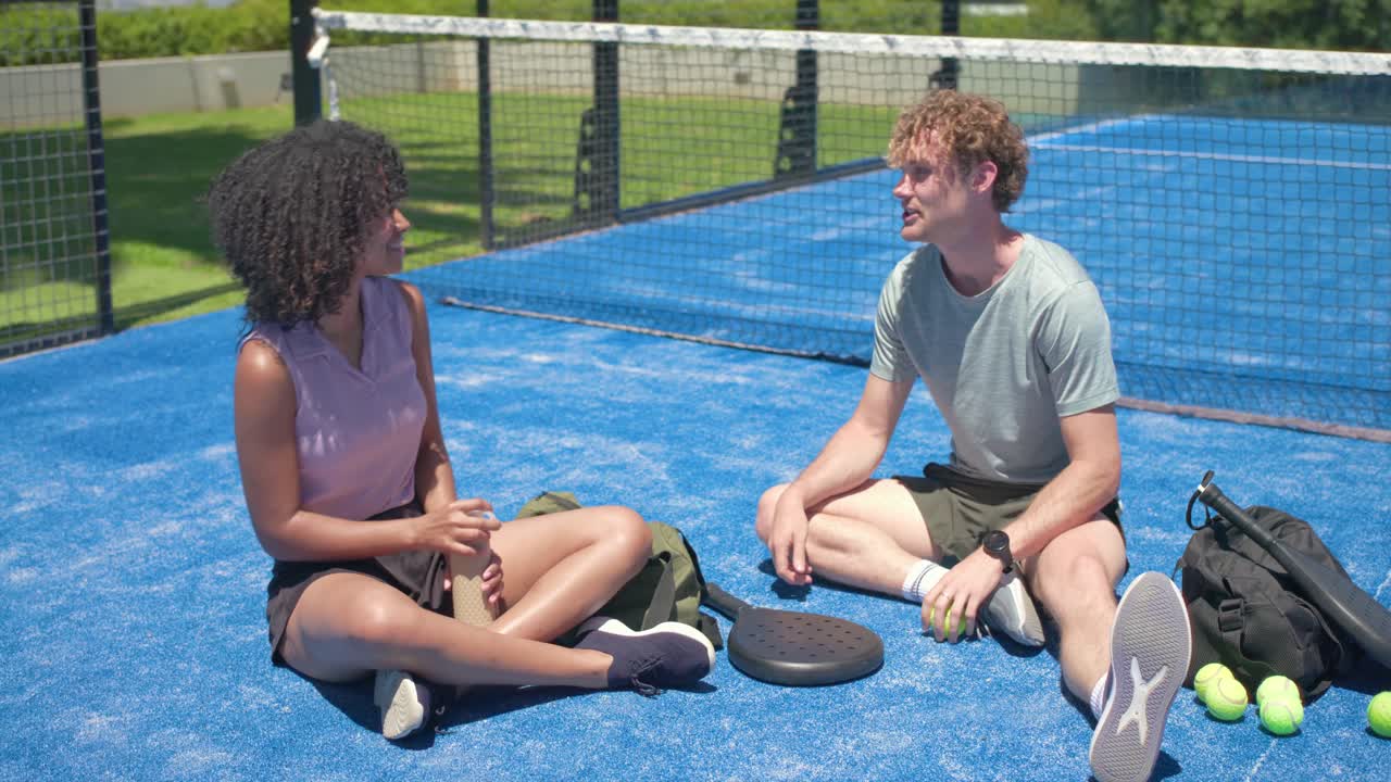 Diverse couple sitting on blue court, man tossing balls by paddles, woman sipping bottle on break