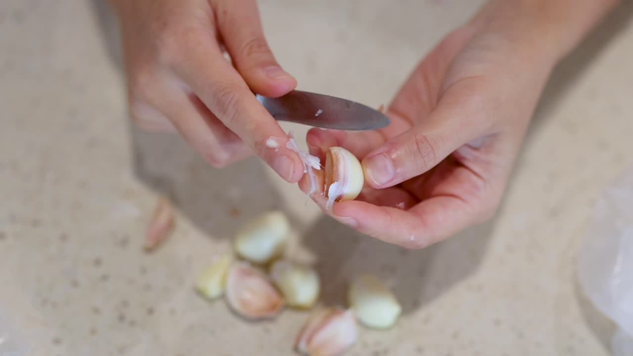 Close-up of hands peeling garlic cloves on a light-colored kitchen surface, using a small knife under bright lighting