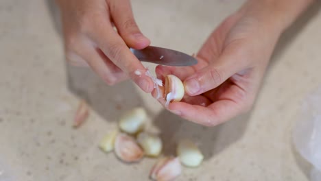 close-up of hands peeling garlic cloves on a light-colored kitchen surface, using a small knife under bright lighting