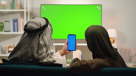 middle eastern couple watches green screen tv while holding a green screen smartphone at home
