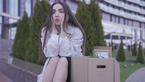 primer plano de una hermosa mujer sentada frente a un edificio de oficinas. ella tiene sus cosas en una caja. ella ha sido despedida. ella está triste.