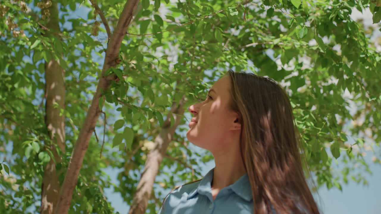 Mujer informal bajo la sombra de hojas, mujer urbana que se detiene y habla por teléfono bajo el follaje, mujer vestida de forma casual conversando por teléfono bajo el vibrante dosel de un árbol en un parque urbano