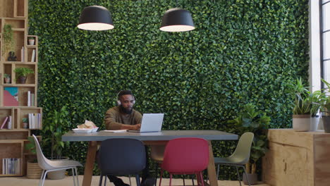 Joven-Hombre-De-Negocios-Afroamericano-Trabajando-Hasta-Tarde-Usando-Una-Computadora-Portátil-Intercambiando-Ideas-Escuchando-Música-Tomando-Café-En-Una-Moderna-Oficina-De-Inicio