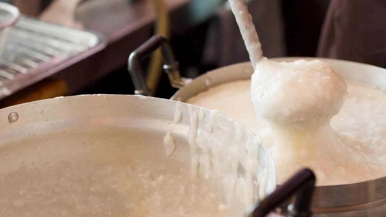 Close-up shot of a ladle stirring thick white rice porridge in a commercial kitchen.