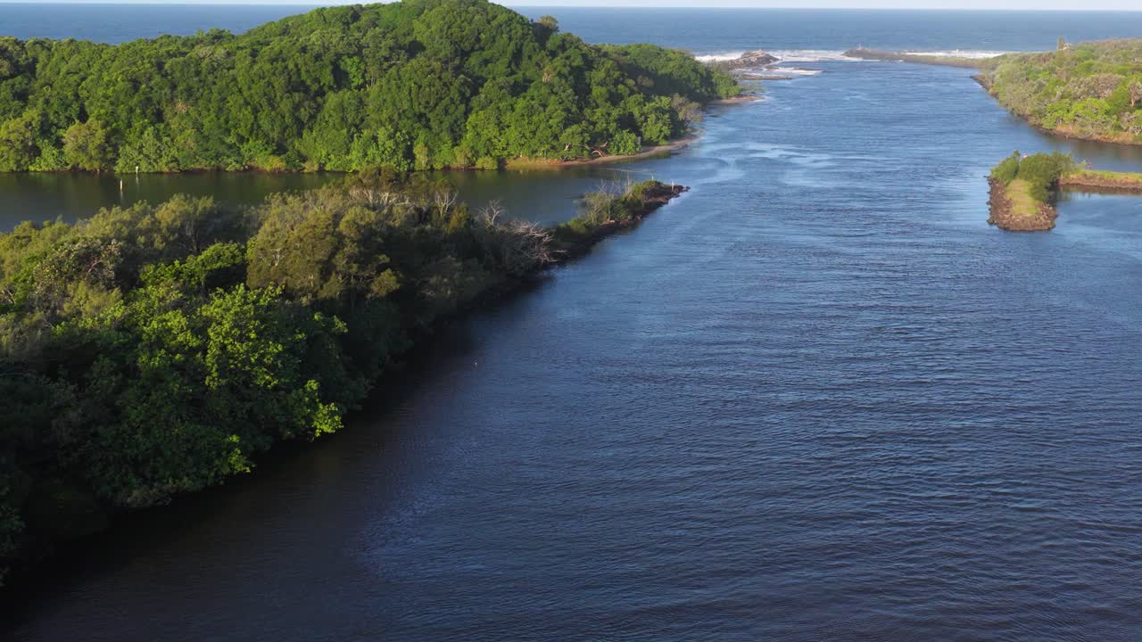 Drone footage captures a serene river flanked by lush greenery under bright daylight in Brunswick Heads, Australia