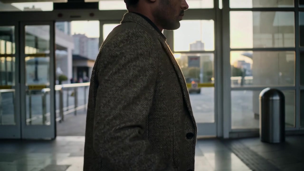 Uomo che cammina in soprabito di tweed, tirando una valigia con ruote, uscendo in piazza con le porte che si aprono.