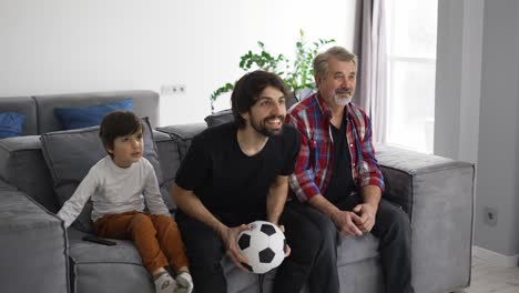 Three-generations-of-men-on-couch-in-living-room-watch-football-match