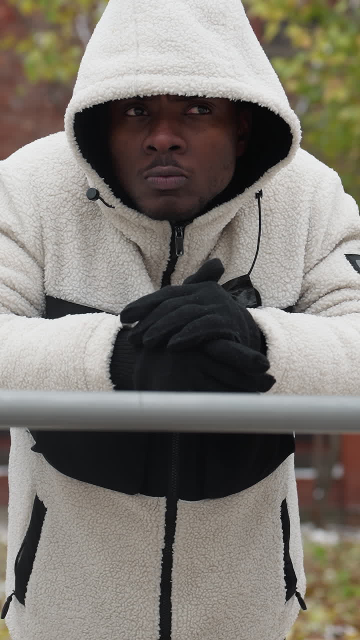 joven rompe los nudillos en una sudadera blanca al aire libre, descansando en una barra de hierro con aire frío visible desde la boca, fondo urbano con edificios, suelo nevado y equipos de entrenamiento de madera