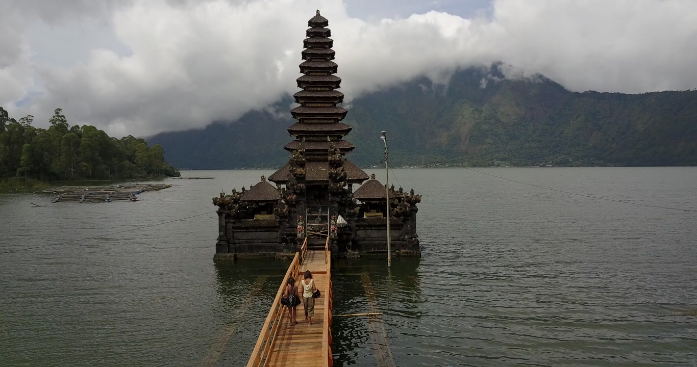 Lifestyle travel clip of people walking along a wooden walkway toward Ulun Danu Beratan Temple, located on Lake Beratan, with Mount Catur and misty clouds in the background in Bali, Indonesia.