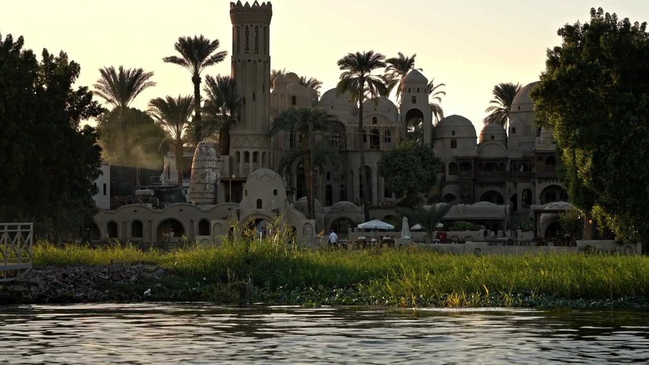 Uno scatto lungo il fiume che cattura una tenuta o un resort ornato, simile a un castello, costruito in stili di design tradizionali egiziani e nubiani lungo la riva del Nilo.
