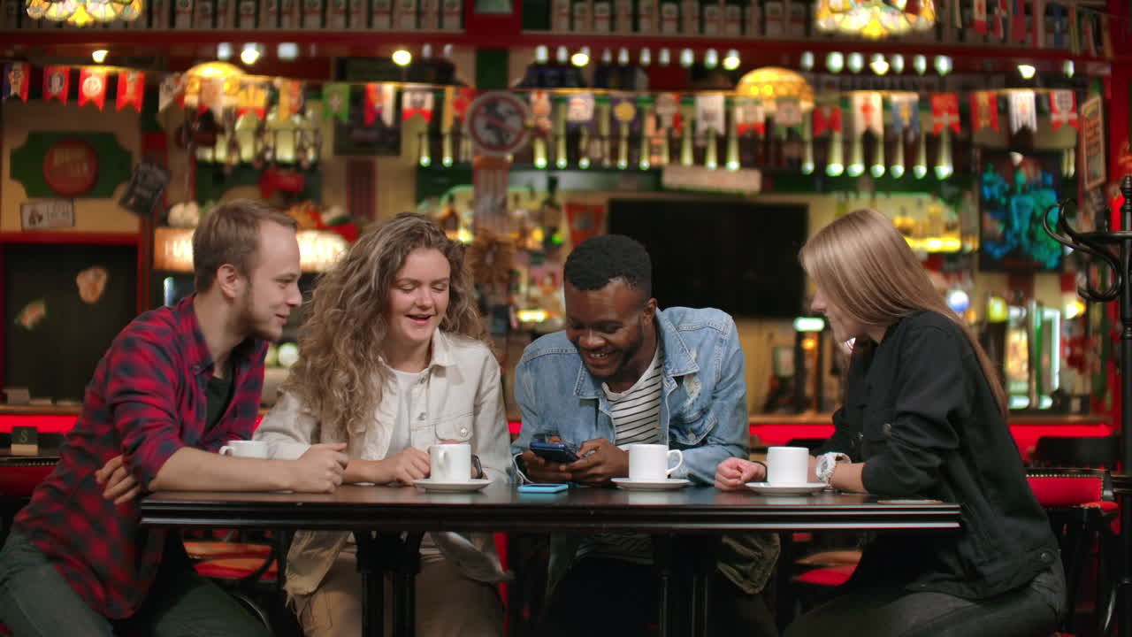 Portrait of cheerful young friends looking at smart phone while sitting in cafe. Mixed race people sitting at a table in restaurant using mobile phone.