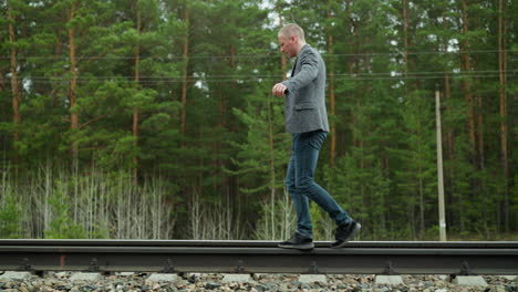 a man in a grey suit jacket and blue jeans walks carefully along a railway track, balancing himself, the background features lush green trees