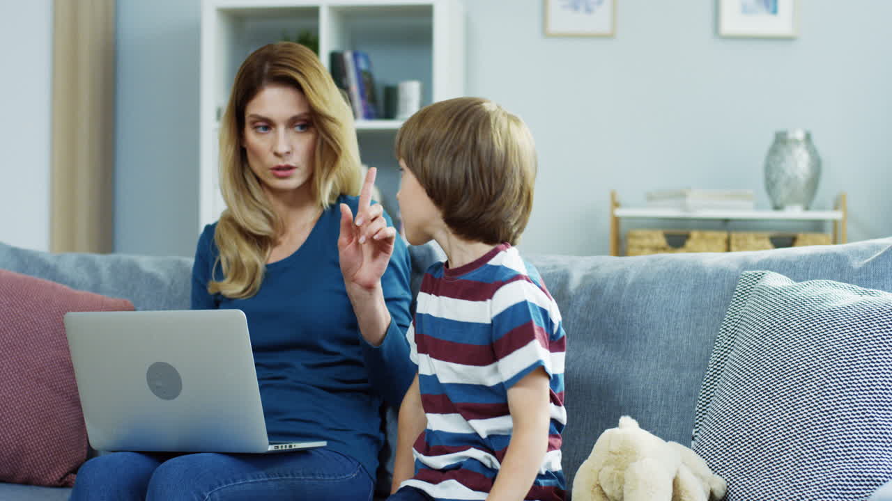Bella madre seduta sul divano a casa con un computer portatile, che vieta qualcosa al figlio piccolo