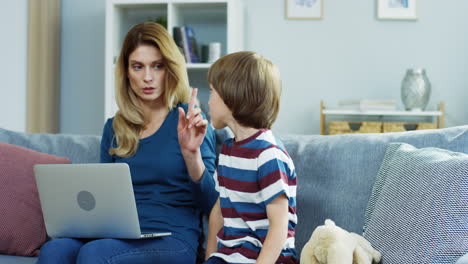 Gut-Aussehende-Mutter,-Die-Zu-Hause-Mit-Einem-Laptop-Auf-Der-Couch-Sitzt-Und-Seinem-Kleinen-Sohn-Etwas-Verbietet