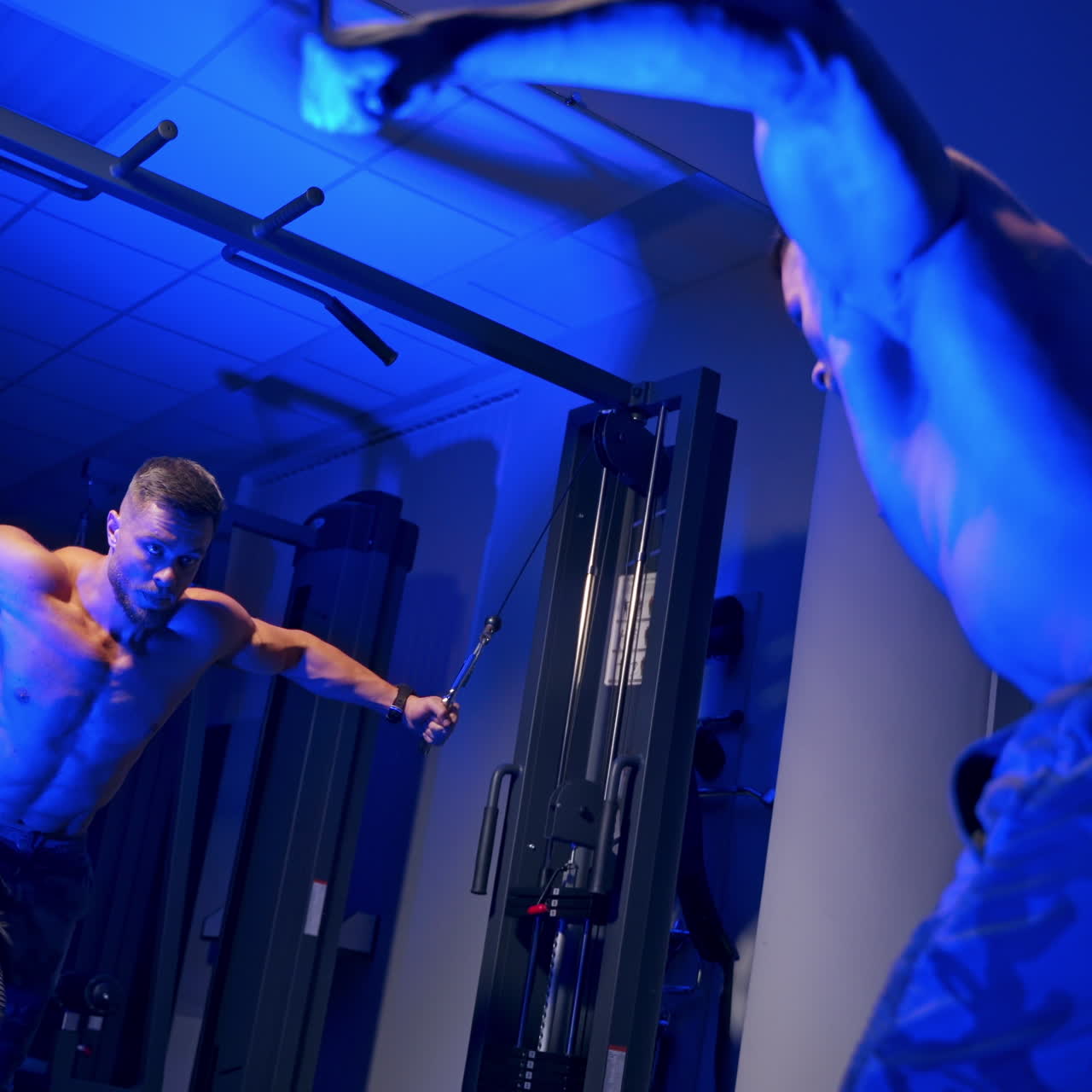 Shirtless bodybuilder during workout in blue light. Reflection in a mirror of a young bodybuilder training his biceps. Muscular man exercising in the gym.