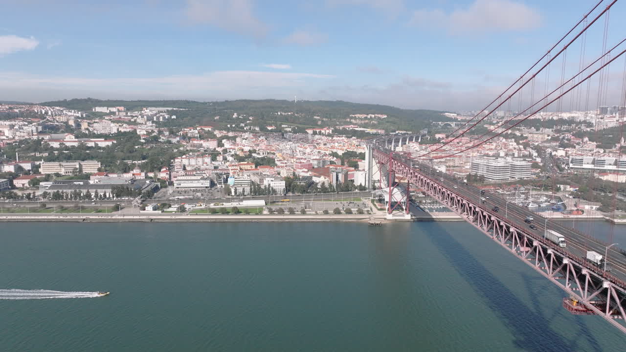 Ripresa aerea con drone del Ponte 25 de Abril a Lisbona, Portogallo, Europa. Soleggiato e luminoso con nuvole sullo sfondo. Auto che guidano sul ponte con barca che va veloce. Girato in ProRes 422 HQ
