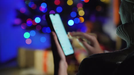 Emale-Hände-Mit-Einem-Smartphone-Auf-Einem-Hintergrund-Des-Weihnachtsbaumlichtergeschenkbestellkonzeptes