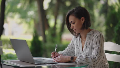 Business-Woman-Brunette-Hispanic-Ethnicity-Works-remotely-while-sitting-in-a-summer-cafe-on-a-sunny-day-with-a-laptop-and-writes-down-with-a-pen-and-notebook