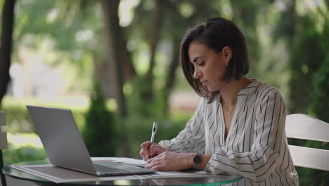 business woman brunette hispanic ethnicity works remotely while sitting in a summer cafe on a sunny day with a laptop and writes down with a pen and notebook