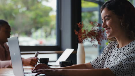 young businesswoman with coffee working on laptop sitting in cafe with colleague in background
