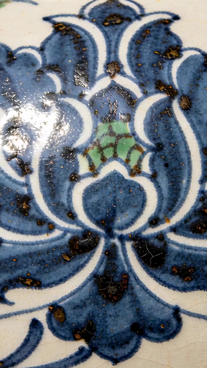 Video verticale: Telecamera in movimento che rivela una piastrella smaltata blu con motivo floreale su una scrivania, mostrando un punto verde e screpolature.