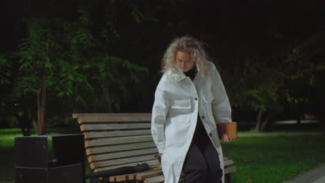 young adult in white coat dropping umbrella on wooden bench at night preparing to sit down and read book under bright streetlights surrounded by greenery