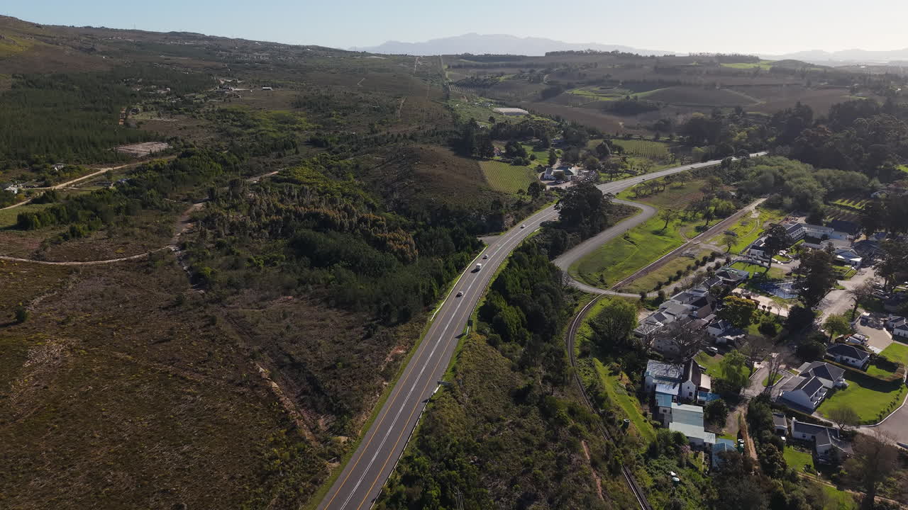 South Africa - A Winding Road Cuts through a Wide Valley Filled with Scattered Homes, Green Fields, and Rolling Hills Under a Bright, Hazy Sky - Aerial Drone Shot