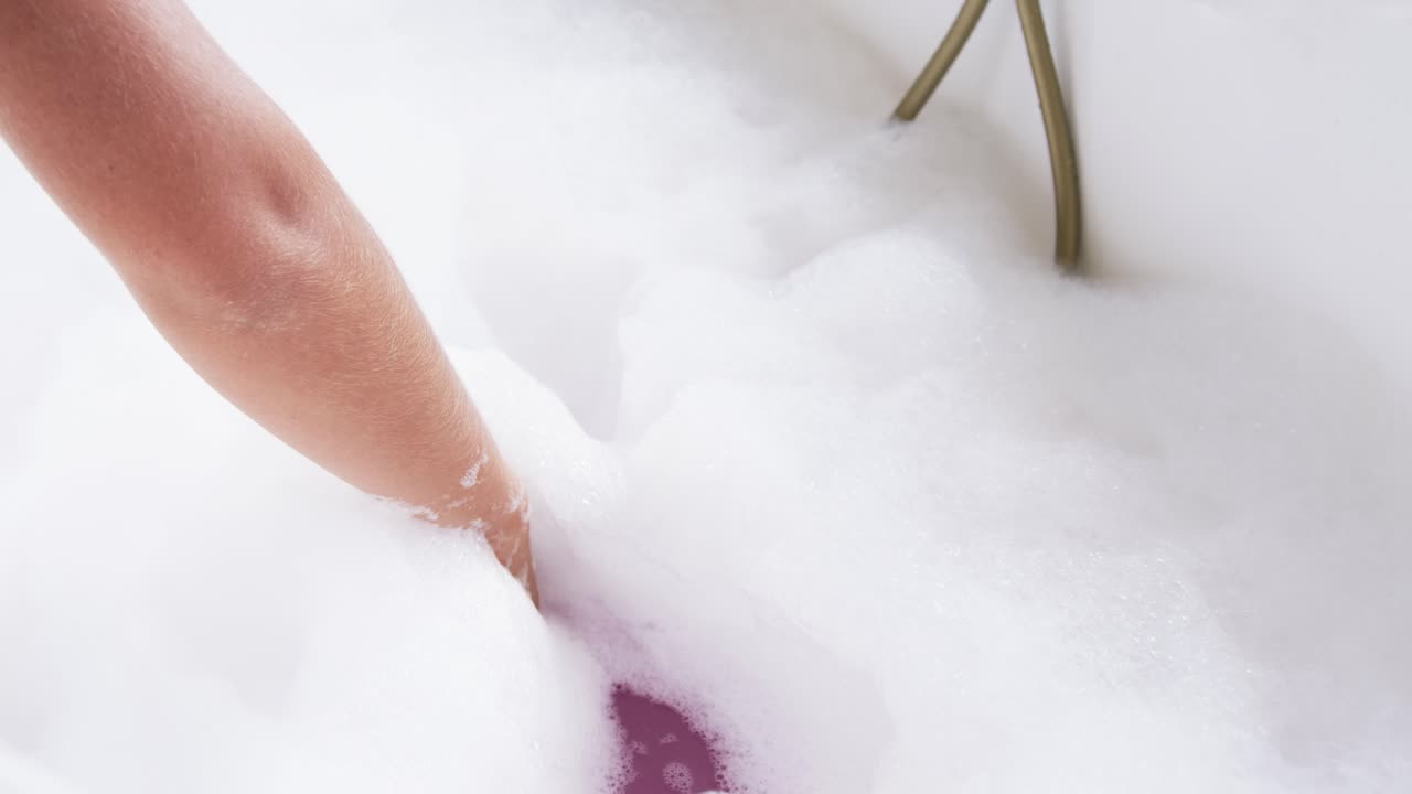 Avambraccio femminile che entra da sinistra spingendo la schiuma nella vasca, rivelando acqua viola e un rubinetto, con spazio per il testo.