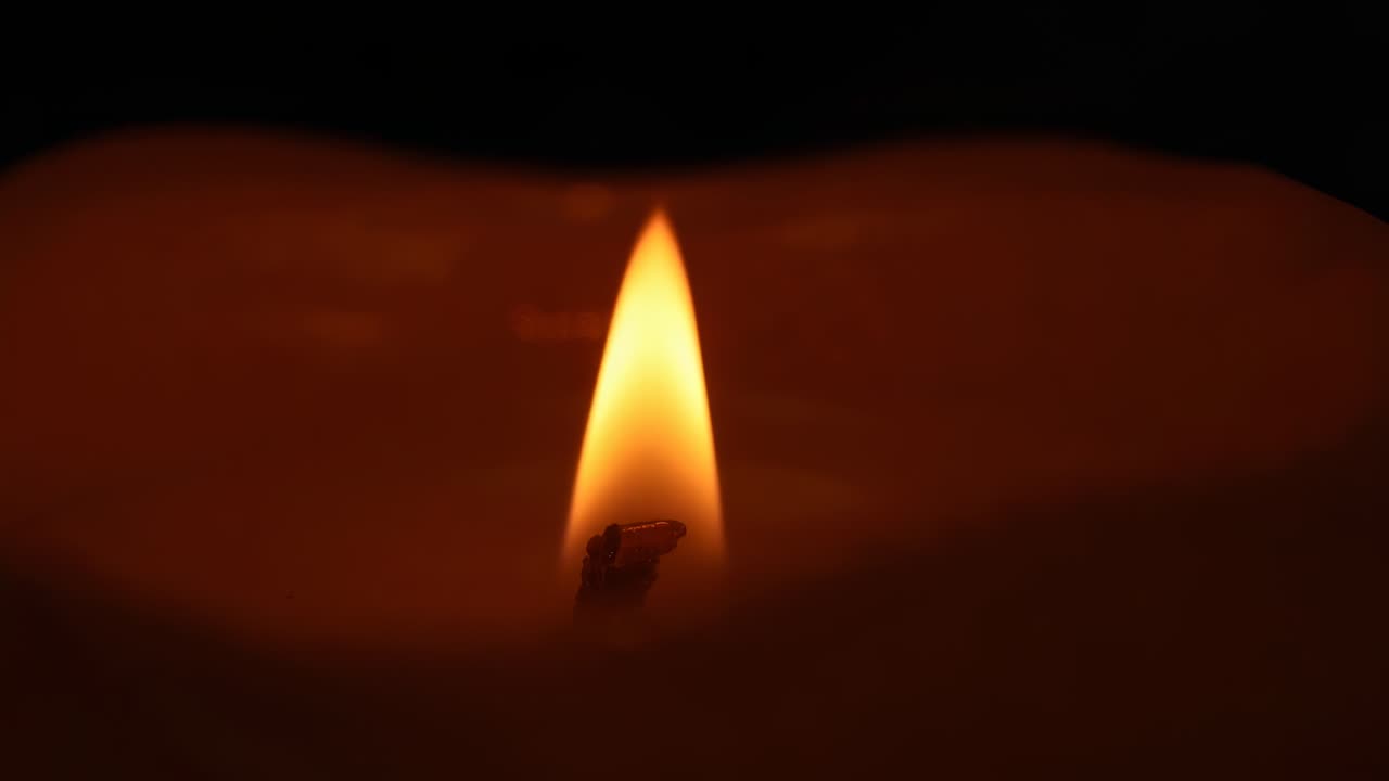 Primi piani su stoppino di candela e cera fusa in una stanza buia, con fiamma giallo-arancione tremolante