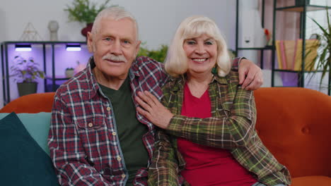 Happy-old-senior-elderly-family-couple-hugging,-laughing,-smiling-looking-at-camera-at-home-sofa