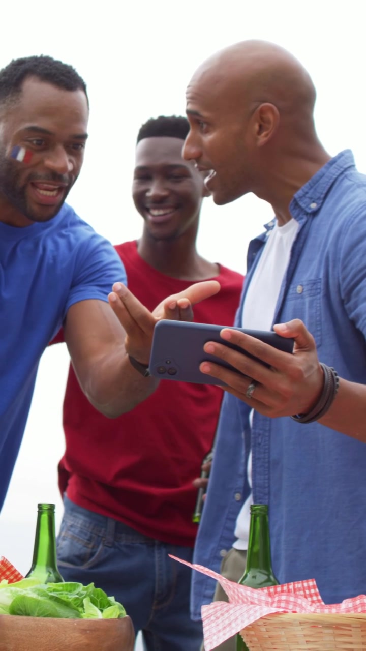Vertical video: Raising bottle, man in red tee leading clap, leaning to view phone by picnic table