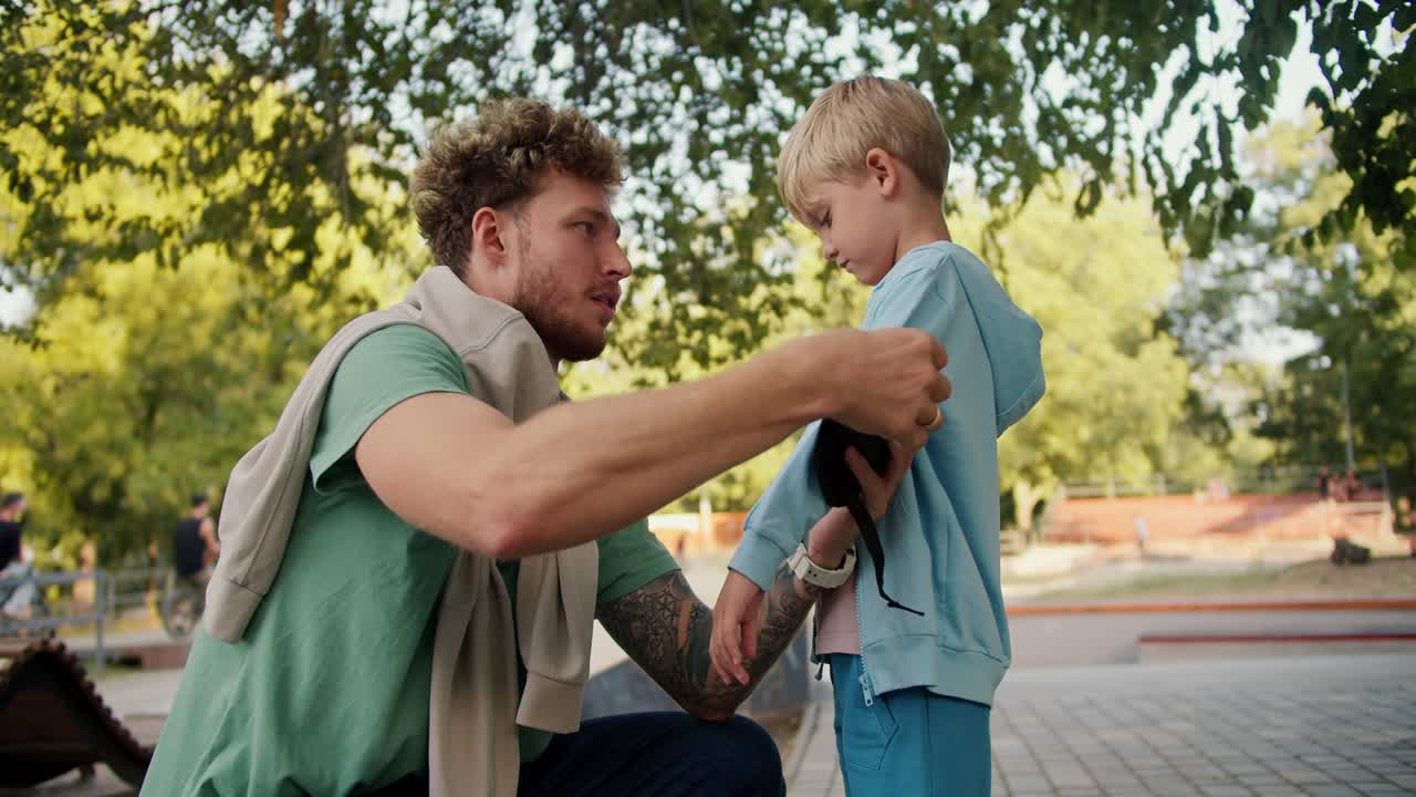 un hombre con cabello rizado y paja en una camiseta verde ayuda a su hijo en un suéter azul a ponerse las almohadillas de los codos antes de montar en una patineta en un parque de patinaje en el parque. un niño rubio pequeño preparándose para montar en un patineta