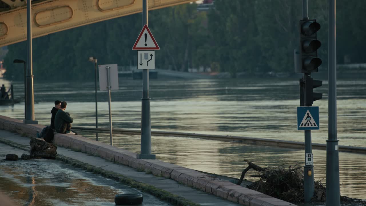 Two people sit near a flooded walkway with submerged traffic signs along the Danube during Budapest Flood 2024