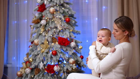 blonde woman in white dress sits on the sofa holding a baby. little kid starts to cry and mom comforts him. christmas tree at backdrop.