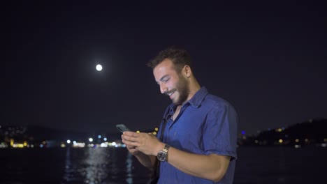 young man using phone at night against city view by the sea.