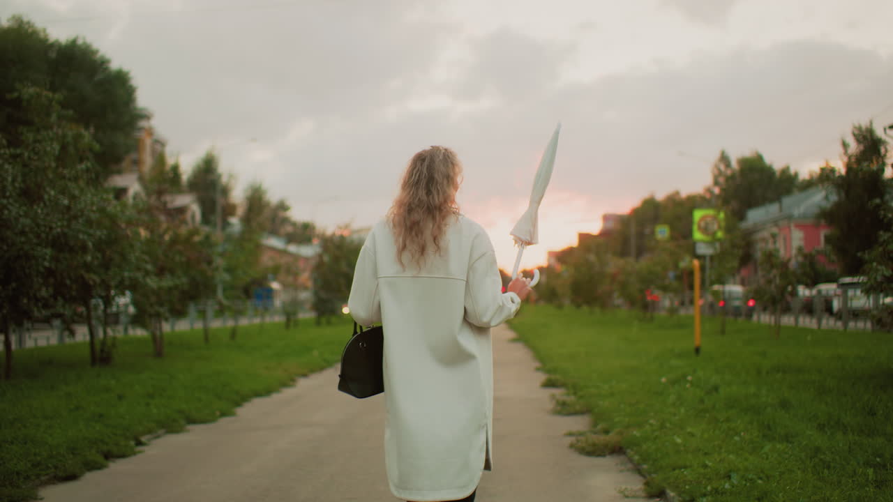 back view of young girl with curly hair walking along paved walkway twirling white umbrella during beautiful sunset in urban park area with green grass and distant cityscape