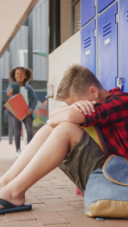 vertical video of sad caucasian boy sitting on floor by lockers at school, copy space