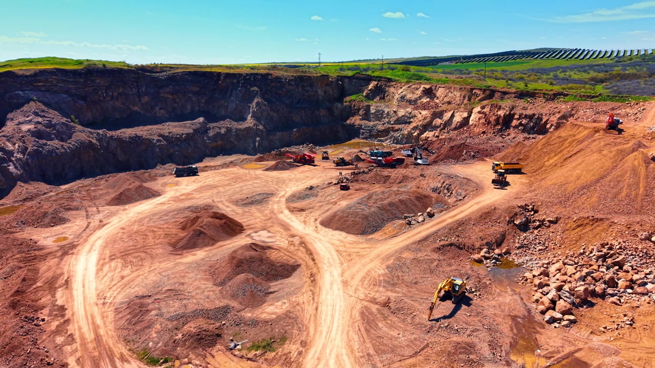 Dagbrudd med tungt maskineri og steinhauger. Bredt luftpanorama av industrielt gruvelandskap med lastebiler og rød jord under blå himmel