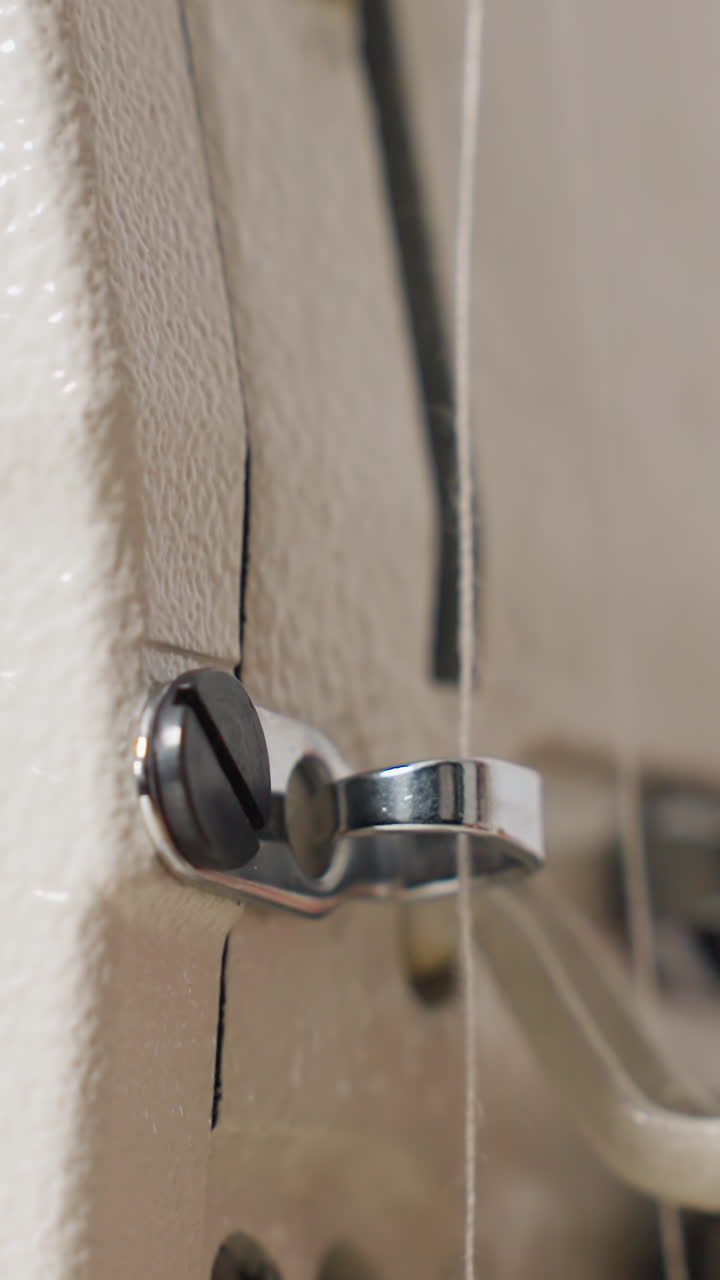 Primer plano de hilo pasando por una guía metálica cerca de la aguja; modista sujetando hilo blanco fino con el pulgar; carcasa texturizada de la máquina con luz suave; mantenimiento rutinario antes de que comience la costura, composición