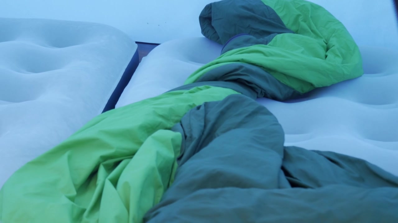 Mão coloca saco de dormir verde sobre dois colchões de ar inflados dentro de uma barraca montada