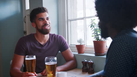 Mixed-race-young-man-and-his-black-dad-raising-beer-glasses-and-talking-sitting-at-a-table-in-a-pub,-close-up,-son-facing-camera