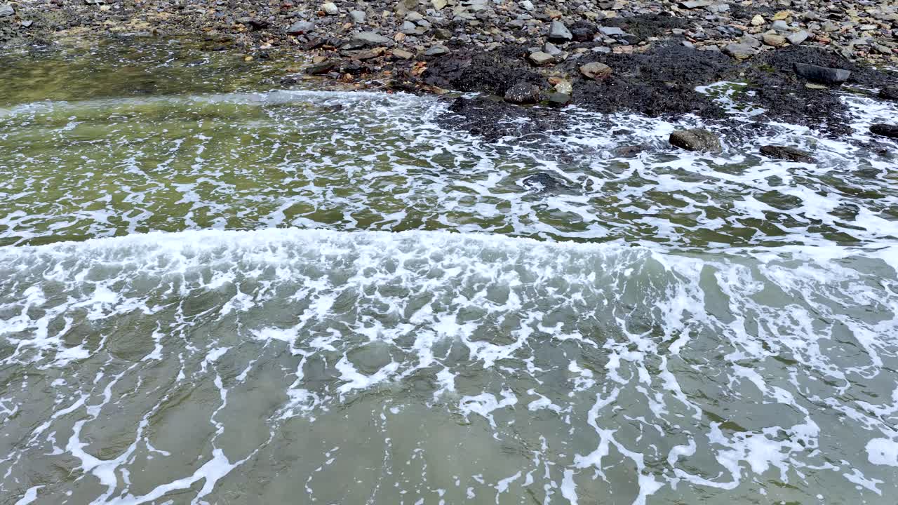 Gentle ocean waves break and recede over rocky shoreline, daylight, static camera, natural soundscape