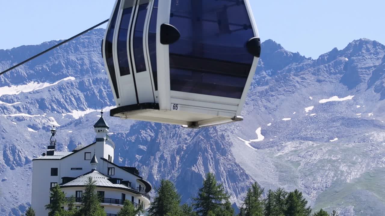 Una funivia bianca scivola accanto a un hotel di montagna, sullo sfondo di cime rocciose