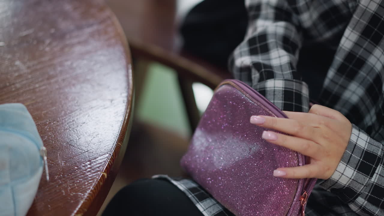 Close-up shot of young lady in checkered shirt opening sparkly purple purse and bringing out cosmetic items on wooden table, well-manicured hands and soft lighting create an elegant beauty