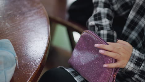 close-up shot of young lady in checkered shirt opening sparkly purple purse and bringing out cosmetic items on wooden table, well-manicured hands and soft lighting create an elegant beauty