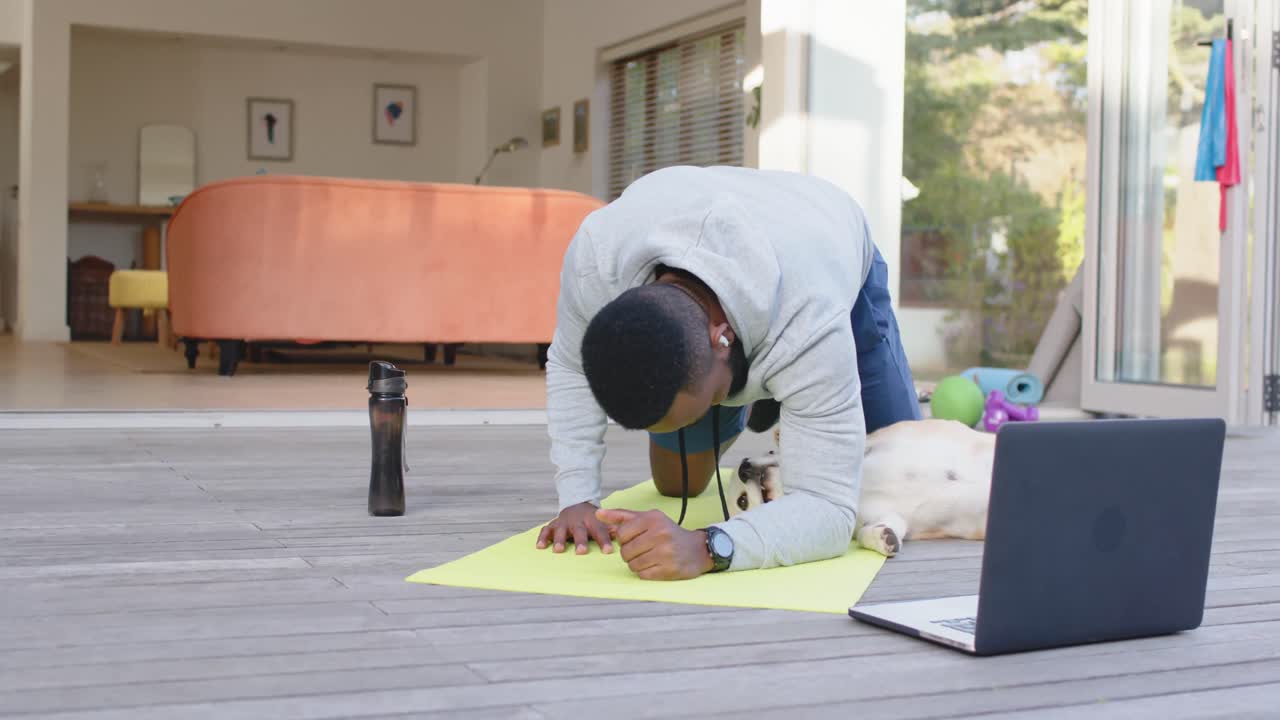 African american man with dog exercising on mat with laptop at home, slow motion