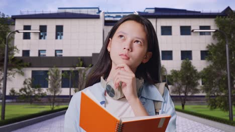 close up of asian teen girl student with a backpack reading book and thinking and looking around then raising her index finger while standing in front of a school building