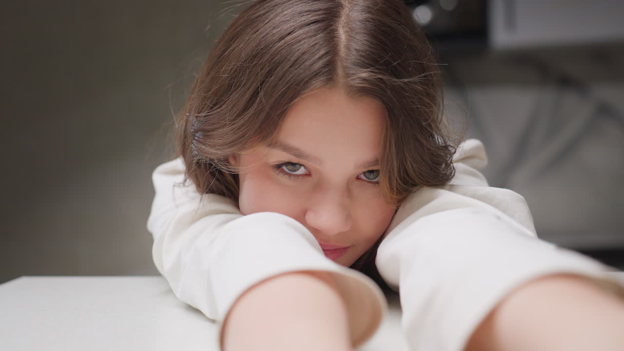Girl seated at table stretches arms forward resting hands flat on surface, casual indoor moment showing relaxation and youthful expression in kitchen environment with soft lighting